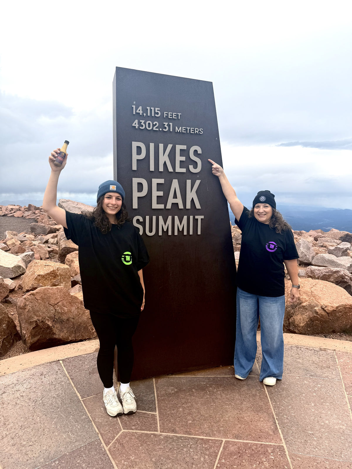Two people in Tranquil Heat shirts holding a hot sauce bottle at the Pikes Peak Summit sign