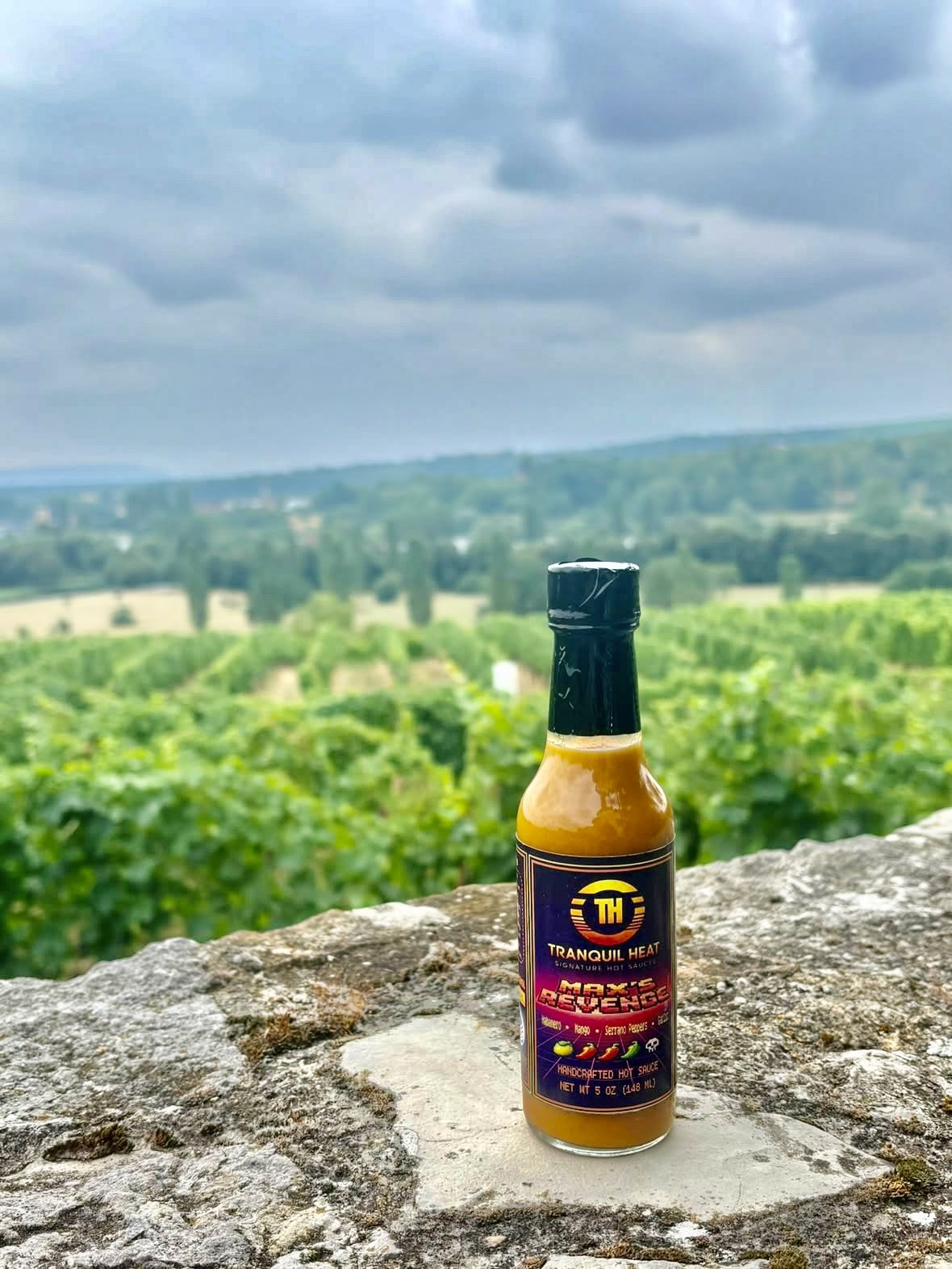 Bottle of Tranquil Heat Max's Revenge hot sauce on a stone wall overlooking a vineyard