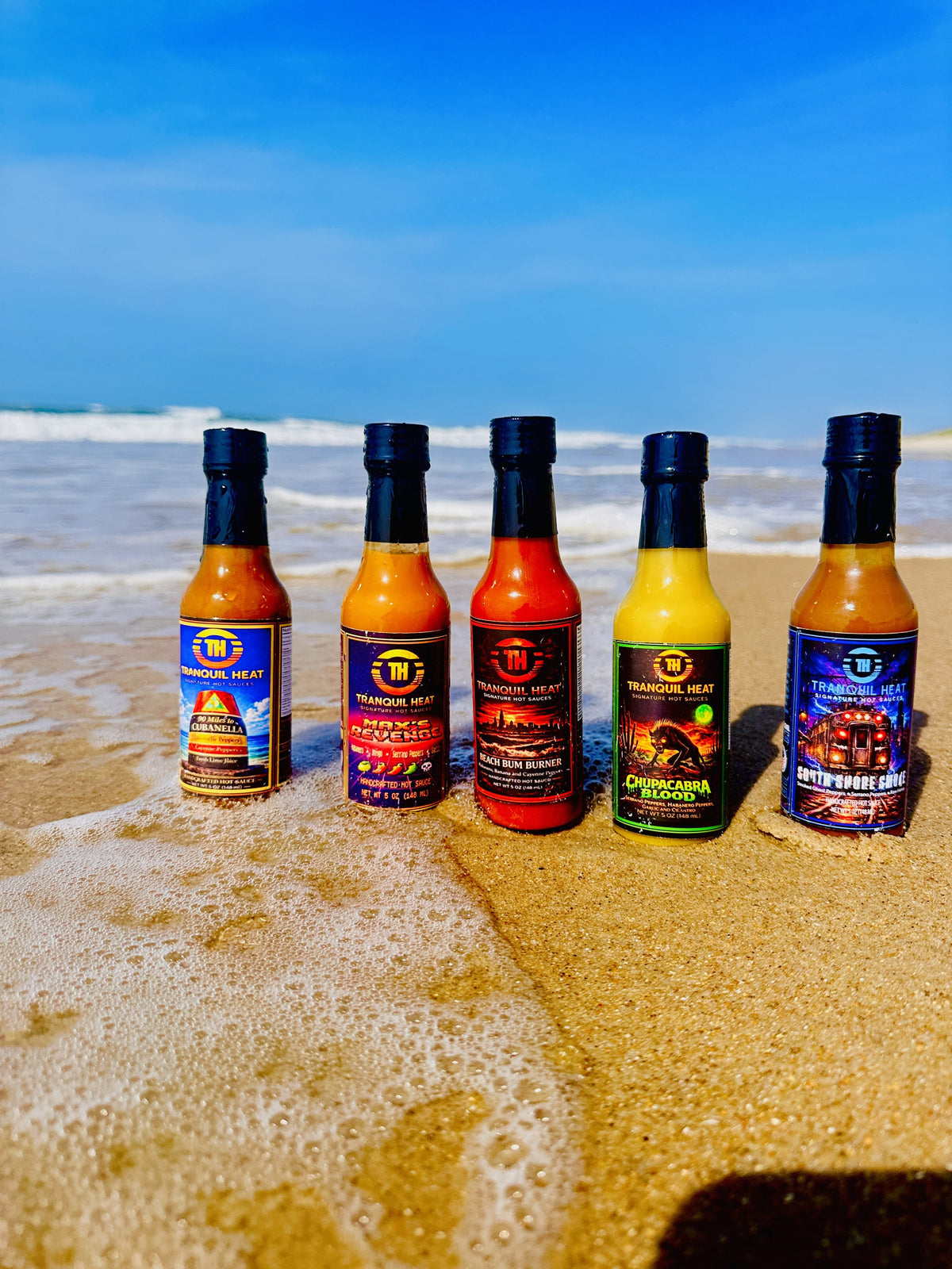 Five Tranquil Heat hot sauce bottles lined up on the beach with ocean waves