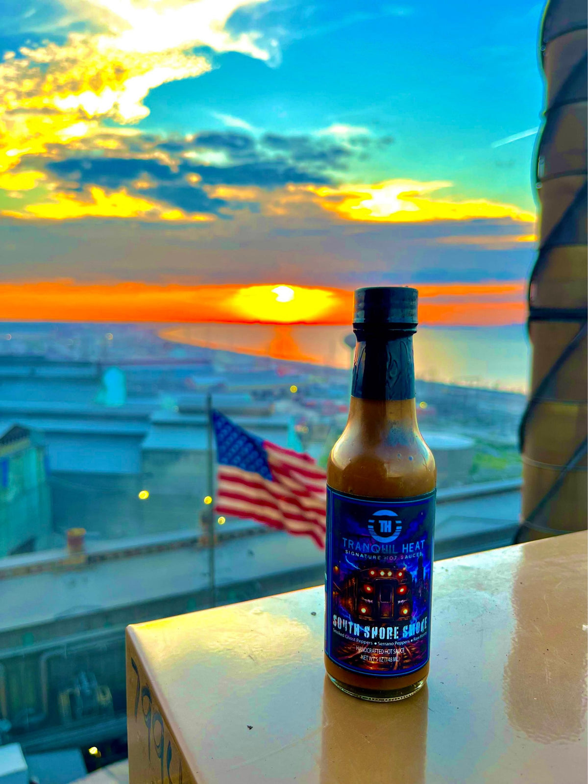 Bottle of Tranquil Heat South Shore Smoke hot sauce at sunset overlooking a lakefront skyline with an American flag