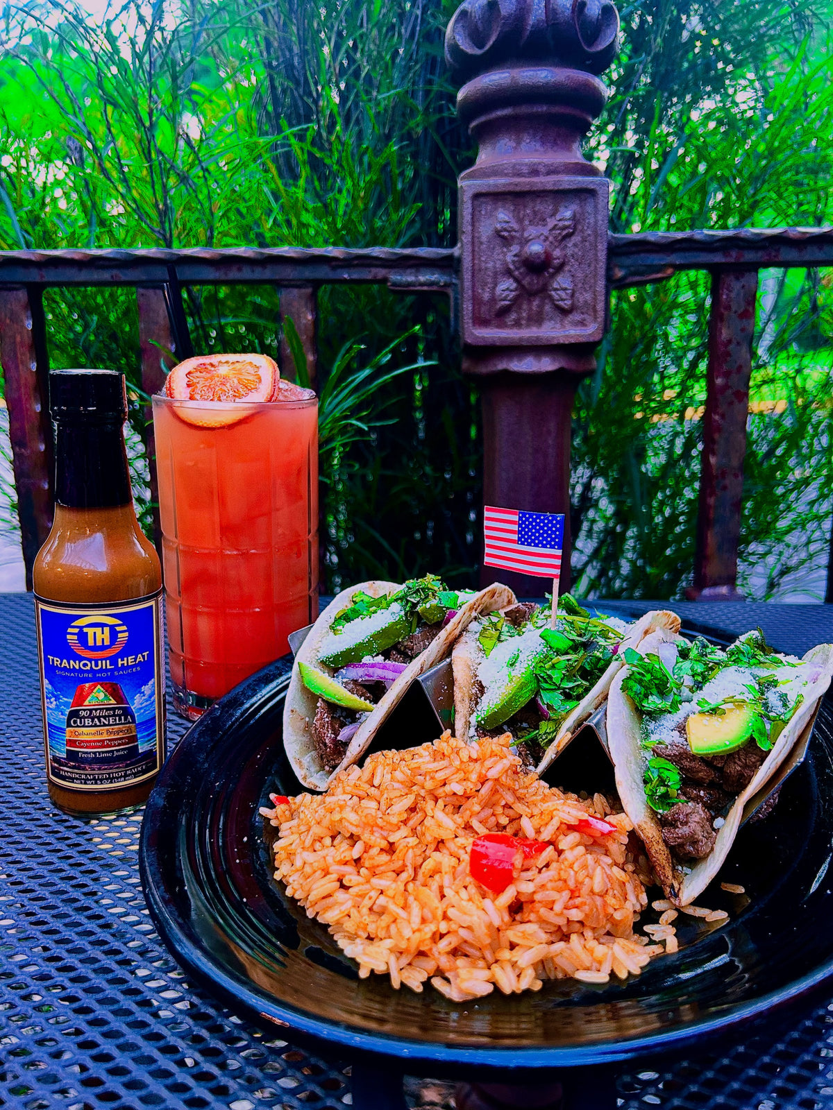 Bottle of Tranquil Heat 90 Miles to Cubanela hot sauce with tacos and a cocktail on a patio table