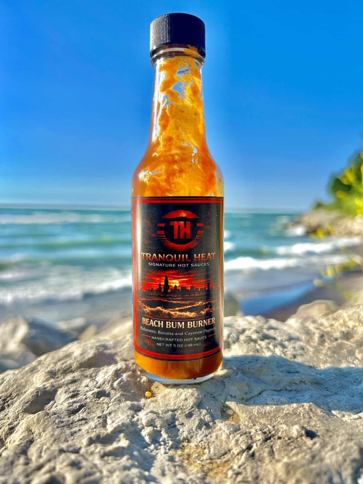 Bottle of Tranquil Heat Beach Bum Burner hot sauce on a rock along the shoreline